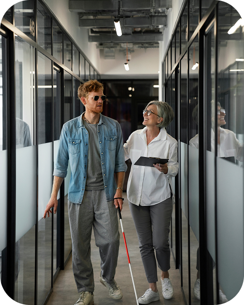 Hombre con discapacidad visual usando bastón blanco siendo guiado por una mujer profesional en un pasillo de oficina moderno