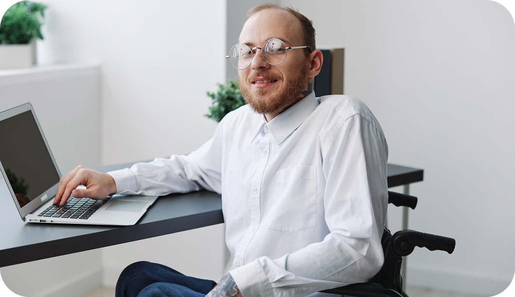 Hombre con discapacidad usando gafas y sentado en una silla de ruedas sonriendo mientras trabaja en un portátil en un escritorio moderno con ambiente luminoso